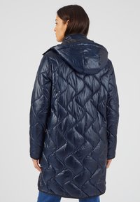 Cappotto imbottito blu navy con un motivo trapuntato, dotato di cappuccio e finitura lucida. Il cappotto arriva a metà coscia, con maniche lunghe e taglio dritto.