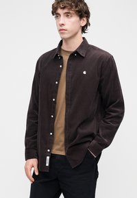 Rjava kordurova srajca z zapenjanjem na gumbe, subtilno teksturo in belim logotipom na prsih, nošena preko rjave majice in črnih hlač.