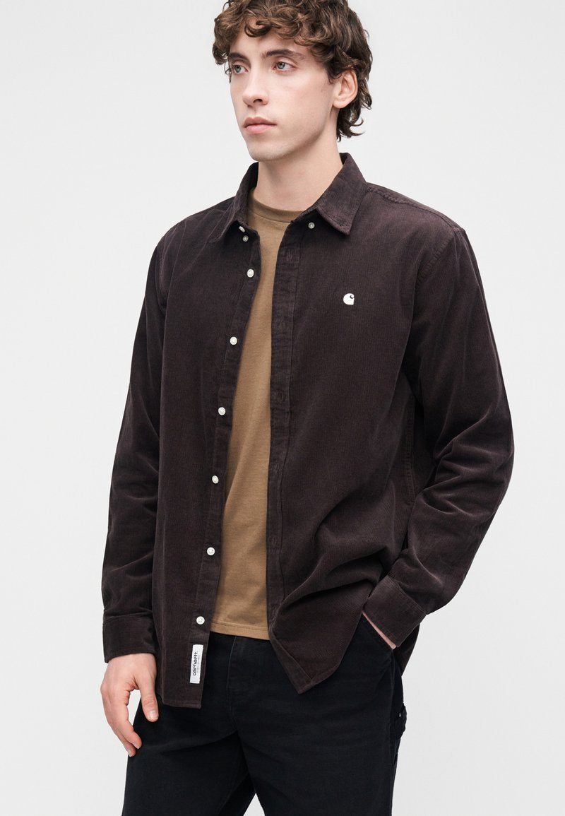 Camicia marrone in velluto a coste con design a bottoni, texture sottile e logo bianco sul petto, indossata sopra una t-shirt marrone e pantaloni neri.