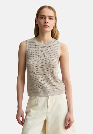 Jeune femme portant un haut en crochet beige sans manches et un pantalon taille haute blanc cassé, debout devant un fond uni clair.