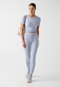Hellblaues, kurz geschnittenes Sportoberteil, kombiniert mit passenden, hoch taillierten Leggings aus strukturiertem Stoff und mit dezentem Logo. Model trägt eine beige Tasche.