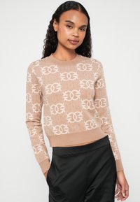 Suéter de malha bege com padrão de logótipos em relevo, decote redondo, mangas longas e bainha curta. Combinado com calças pretas.