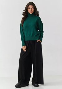 Zielony sweter z golfem wykonany z dzianiny, z prążkowanym wykończeniem, zestawiony z luźnymi czarnymi spodniami o szerokich nogawkach i czarnymi sneakerami.