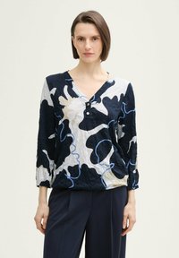 Blouse froissée avec un col en V et des accents de boutons. La palette de couleurs comprend des motifs abstraits en bleu marine, crème et bleu clair. Manches longues.