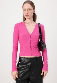 Pink long-sleeve top with a V-neck, featuring ruching details and metallic buttons, paired with black faux leather pants and a small handbag.