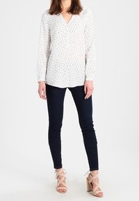 Blusa bianca leggera con piccole pois nere, scollo a V, maniche lunghe, abbinata a jeans skinny scuri e sandali con tacco alto beige.