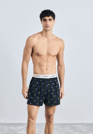 Boxers em azul escuro com riscas verticais. Apresenta uma cinta elástica branca com o logótipo da Lacoste e um padrão de pequenos crocodilos.