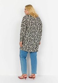 Tunika lunga nera e bianca con un motivo, maniche lunghe, abbinata a jeans blu chiaro e sandali con tacco rossi. Si nota il design del tessuto testurizzato.
