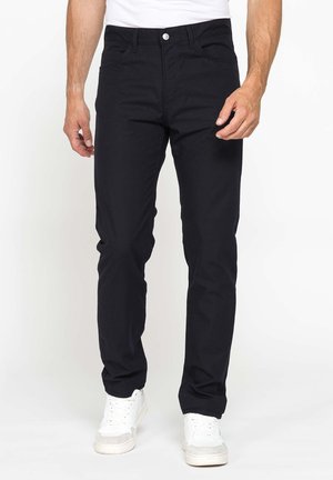 Uomo che indossa jeans neri slim fit e sneakers bianche, in piedi su uno sfondo bianco uniforme.