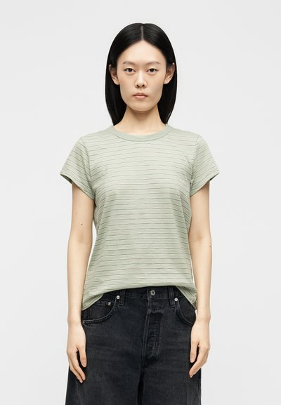 Γυναίκα με ίσια μαύρα μαλλιά, που φοράει ένα ανοιχτό πράσινο ριγωτό κοντομάνικο μπλουζάκι και μαύρα τζιν ψηλόμεσα, στέκεται μπροστά σε απλό φόντο.