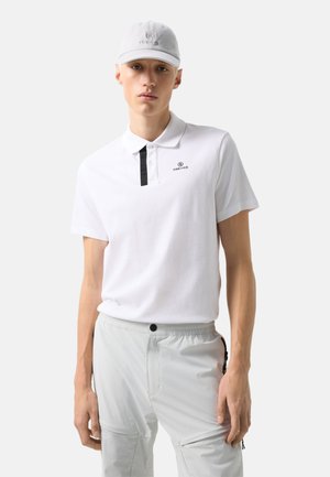 Młody mężczyzna ubrany w białą koszulkę polo Fire + Ice, pasującą białą czapkę i jasnoszare spodnie z zapinanym na guziki pasem.