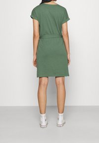 Oliwkowa sukienka z krótkim rękawem, z wcięciem w talii, o prostym kroju i miękkiej fakturze, zestawiona z białymi sportowymi trampkami.