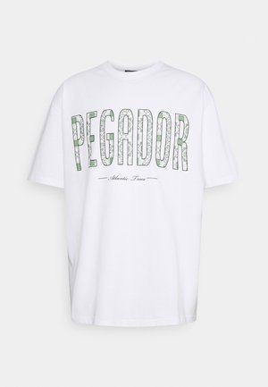 Camiseta blanca de algodón con la palabra "PEGADOR" en verde y negro con un patrón decorativo, y "Atlantic Trace" debajo en texto más pequeño.