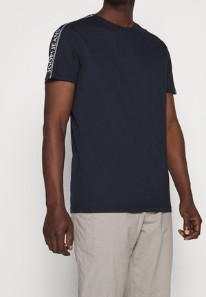 Mand iført en navyblå kortærmet T-shirt med teksten "JOOP! JEANS" på skulderstriberne og lyse beige bukser mod en ensfarvet baggrund.