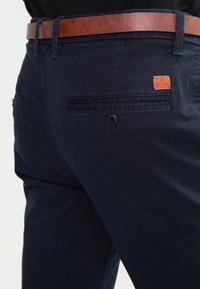 Mörkblå bomullshängselbyxor med ett brunt läderbälte, bakficka med knappstängning och en liten orange märkesetikett.