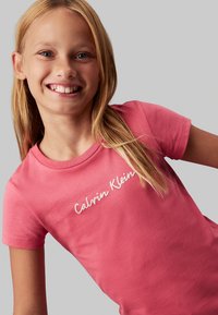 Camiseta de algodón rosa con manga corta, que presenta un logo blanco "Calvin Klein" impreso en diagonal en el pecho. Textura suave y corte clásico.