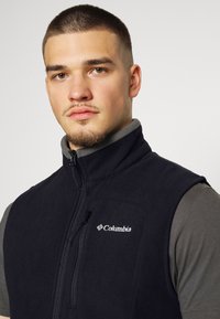 Schwarze Fleece-Weste mit hohem Kragen und Reißverschluss vorne, ausgestattet mit einer seitlichen Tasche und dem Columbia-Logo auf der Brust.
