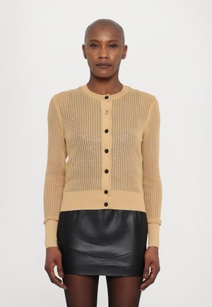 Beige knitted cardigan with vertical ribbing, round neckline, and black buttons. Paired with a black leather mini skirt.