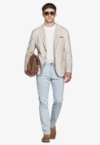 Uomo con occhiali da sole, blazer beige sopra camicia bianca, jeans azzurri, scarpe marroni, che tiene una borsa marrone in una mano.