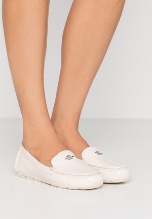 Fødder iført hvide læder slip-on loafers fra Coach med syninger og logo på toppen, stående på en ensfarvet lys overflade.