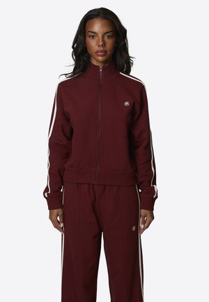 Donna che indossa una giacca bordeaux con zip e pantaloni abbinati con strisce laterali bianche, in piedi rivolta verso la fotocamera su uno sfondo semplice.