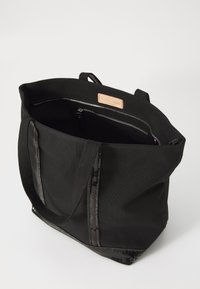 Sac fourre-tout noir en toile avec une texture lisse, deux poignées et une poche intérieure zippée avec un détail en cuir.