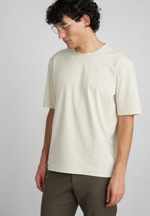 T-shirt de cor creme, de manga curta, feita de tecido macio, com um corte descontraído e um decote redondo, combinada com calças castanhas.
