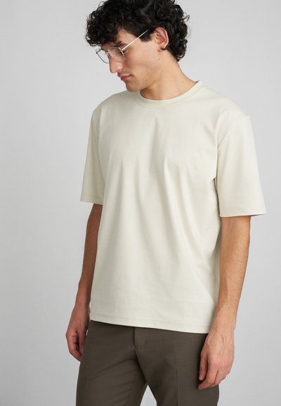 Cremefarbenes T-Shirt mit kurzen Ärmeln aus weichem Stoff, in lässigem Schnitt und Rundhalsausschnitt, kombiniert mit braunen Hosen.