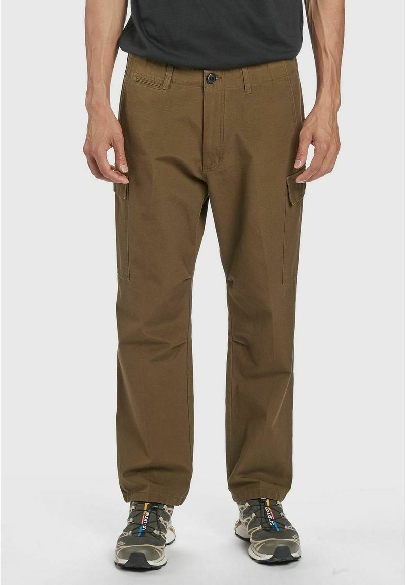 Pantaloni cargo marroni con tasche laterali, taglio dritto, materiale misto cotone e chiusura con bottone. Abbinati a scarpe da trekking grigie e nere.