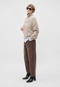 Světle béžový pletený rolák, hnědé široké kalhoty a černé kotníkové boty na podpatku. Doplňky zahrnují hnědé sluneční brýle.