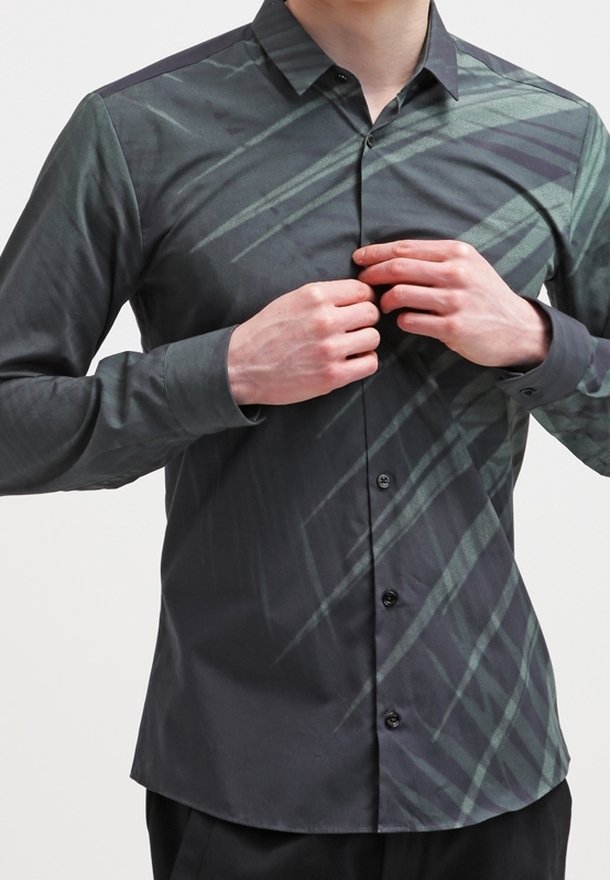 Uomo che allaccia i bottoni su una camicia grigio scuro con un motivo a strisce diagonali verde chiaro, indossando pantaloni neri.
