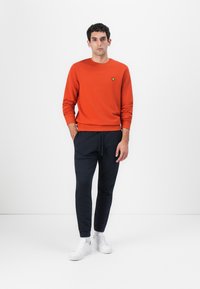 Narancsszínű pulóver kerek nyakkivágással és bordázott mandzsettákkal, fekete, slim-fit melegítőnadrággal és fehér sportcipővel párosítva. Egyszerű dizájn, minta nélkül.