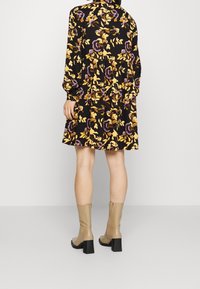 Mujer vistiendo un vestido negro con flores amarillas y moradas, combinado con botas de tobillo de tacón beige, de pie contra un fondo simple.