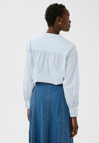 Donna con capelli corti e ricci che indossa una blusa a maniche lunghe azzurro chiaro infilata in una gonna di jeans a vita alta, di spalle contro uno sfondo semplice.