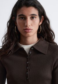 Brown ribbed knit top with a buttoned placket, featuring a collared neckline. Smooth texture with subtle vertical lines and black buttons.