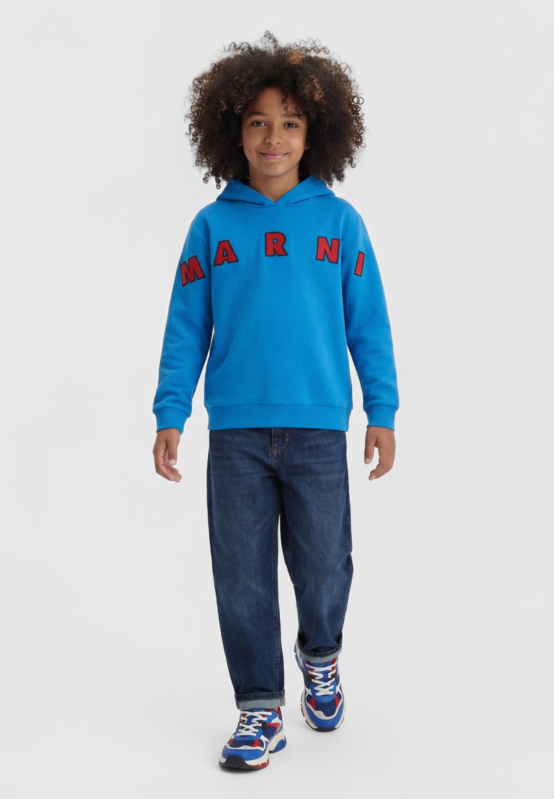 Dziecko z kręconymi włosami w niebieskiej bluzie z kapturem z czerwonymi literami, ciemnych dżinsach oraz niebiesko-biało-czerwonych trampkach idące do przodu na białym tle.