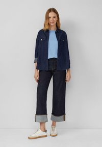 Donkerblauw denim shirt met knopen en twee voorzakken. Lichtblauwe ribbed top eronder. Donkere wijde jeans met oprolmanchetten. Witte sneakers.