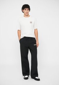 Λευκό κοντομάνικο μπλουζάκι με στρογγυλή λαιμόκοψη και μικρό καφέ λογότυπο; μαύρο παντελόνι φαρδύ με "Double" σε λευκό; συνδυασμένο με μαύρα και άσπρα παπούτσια.