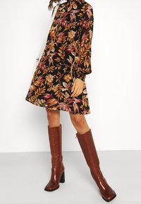 Musta pitkähihaisinen mekko, jossa on oranssit ja vaaleanpunaiset kukkakuosit, yhdistettynä polveen asti ulottuviin ruskeisiin korkokenkiin ja valkoiseen laukkuun.