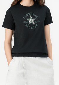 Persona vistiendo una camiseta negra de Converse All Star con el logo de estrella floral y pantalones claros, con las manos en los bolsillos.