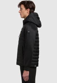 Zwart pufferjack met een afneembare capuchon, gestructureerd gewatteerd lichaam, gladde mouwen, zijzakken en een logo-accent op de linkerschouder.