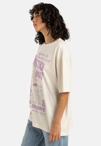T-shirt overdimensionato color crema con stampa grafica viola raffigurante una scena di spiaggia e testo. Maniche corte e scollatura rotonda.