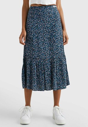 Mujer con una falda midi azul con estampado floral y bajo con volantes, y zapatillas blancas, de pie frente a un fondo claro y liso.
