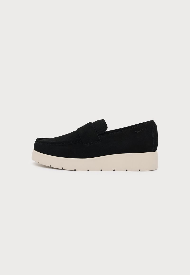 Μαύρο μοκασίνι slip-on με παχιά λευκή σόλα, διαθέτει ευρύ λουράκι μπροστά και διακριτική σήμανση Calvin Klein στη φτέρνα.