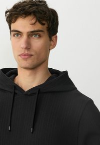 Hanorac negru cu textură canelată, glugă ajustabilă cu șnur și accesorii subtile din metal argintiu. Modelul are o expresie feței curată și neutră.