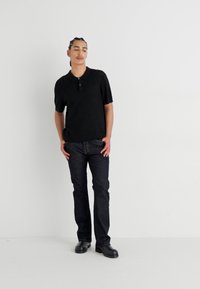 Camisa polo preta de manga curta com colarinho abotoado, combinada com jeans escuros de ganga e sapatos pretos, de pé contra um fundo branco simples.
