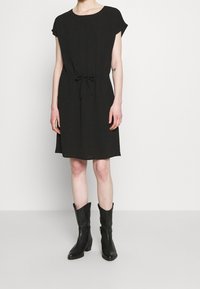 Mujer viste un vestido negro de manga corta con la cintura atada y botas de cuero negro hasta la mitad de la pantorrilla, de pie frente a un fondo blanco.