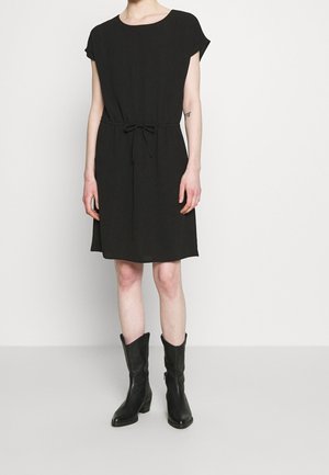 Eine Frau trägt ein kurzärmliges schwarzes Kleid mit einer gebundenen Taille und halbhohen schwarzen Lederstiefeln. Sie steht vor einem weißen Hintergrund.