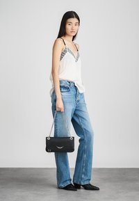 Czarna skórzana torebka na łańcuszku, z srebrnymi akcentami i tłoczeniami. Modelka ubrana w białą bluzkę i niebieskie dżinsy, stojąca na jasnym tle.