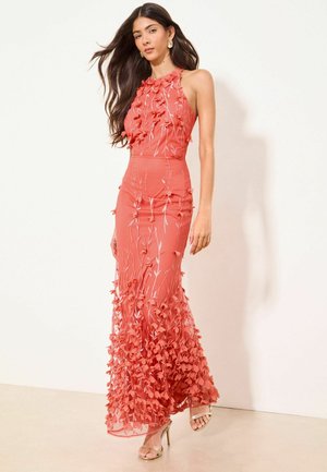 Frau mit langen dunklen Haaren, die ein ärmelloses Korallkleid mit 3D-Blumenapplikationen und goldene High-Heel-Sandalen trägt, steht vor weißem Hintergrund.